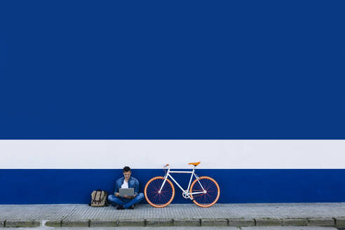Mann in Freizeitkleidung mit Laptop auf Gehweg mit Fahrrad gegen blaue Wand - XLGF01188