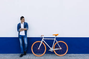 Man in denim casuals with arms crossed standing by bicycle against white wall - XLGF01189