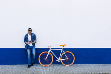 Lächelnder reifer Mann mit Mobiltelefon, der Musik über drahtlose Kopfhörer hört, auf dem Fahrrad an der Wand - XLGF01190