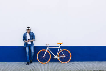 Smiling mature man with mobile phone listening music through wireless headphones by bicycle against wall - XLGF01190