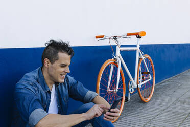 Lächelnder gutaussehender Mann, der sein Mobiltelefon mit dem Fahrrad an der Wand benutzt - XLGF01193