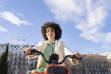 Smiling woman sitting on electric bicycle against cloudy sky - JCCMF01210