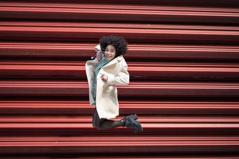 Glückliche Afro-Frau, die gegen einen roten Fensterladen springt - JCCMF01234