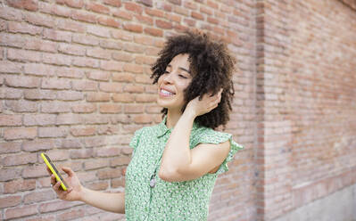 Lächelnde Frau mit Hand in den Haaren, die ihr Smartphone an einer Backsteinmauer hält - JCCMF01236