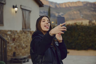 Verspielte Frau streckt die Zunge heraus, während sie ein Selfie mit dem Handy vor einem Haus macht - ACPF01145