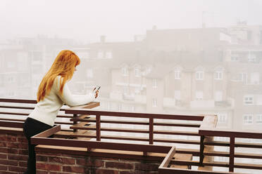 Rothaarige Frau, die ein Mobiltelefon benutzt, während sie sich auf der Terrasse an ein Geländer lehnt - LJF02033