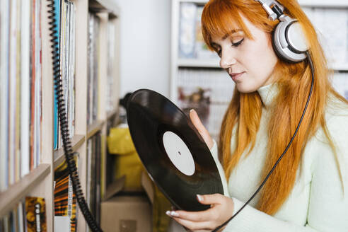 Rothaarige Frau mit Kopfhörern hört Musik und hält eine Schallplatte in einem Geschäft - LJF02047