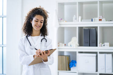 Lächelnde Forscherin schreibt auf einem Klemmbrett, während sie in einer Klinik arbeitet - GIOF11232