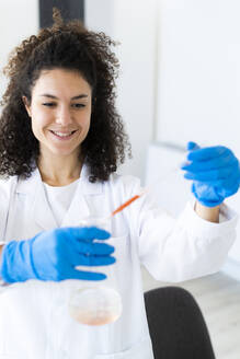 Smiling female doctor mixing solution in beaker through pipette at chemistry lab - GIOF11278