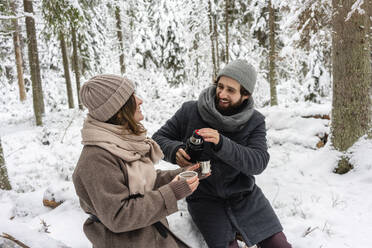 Young couple smiling while having coffee in forest during winter - VPIF03539