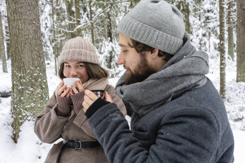 Couple drinking coffee while sitting in forest during winter - VPIF03540