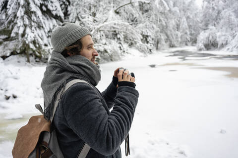 Junger Mann mit Rucksack und Kamera schaut weg, während er im Wald steht, lizenzfreies Stockfoto