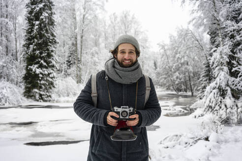 Junger Mann mit Kamera, der lächelnd im verschneiten Wald steht - VPIF03563