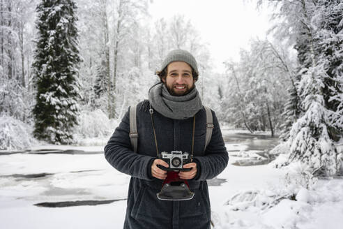 Young man with camera smiling while standing at snowy forest - VPIF03563