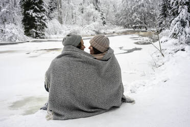 Junges Paar in eine Decke gehüllt, das sich gegenseitig ansieht, während es am gefrorenen Fluss im Wald sitzt - VPIF03564
