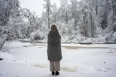 Woman covered in blanket looking at frozen river while standing in forest - VPIF03568