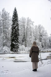 Young woman wearing warm clothing standing by frozen river in forest - VPIF03570