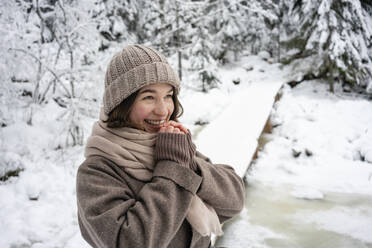 Happy woman wearing knit hat and jacket looking away while standing in forest - VPIF03573