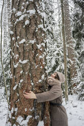 Young woman hugging tree while standing in forest during winter - VPIF03574