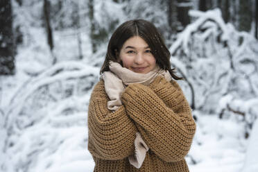 Young woman wearing sweater smiling while standing with arms crossed in forest - VPIF03577