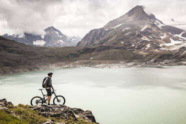 Mountain biker looking at view of Griessee lake while standing at Nufenenpass, Valais, Switzerland - MSUF00503