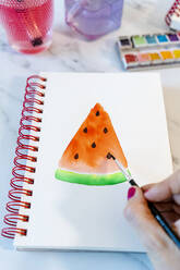 Frau malt Wassermelonenscheibe mit Aquarellfarbe in Buch - GEMF04687
