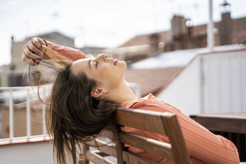 Woman playing with hair while relaxing on chair at rooftop - AFVF08163