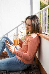 Lächelnde Frau, die einen Smoothie trinkt und dabei ihr Handy benutzt, auf einem Dach sitzend - AFVF08166