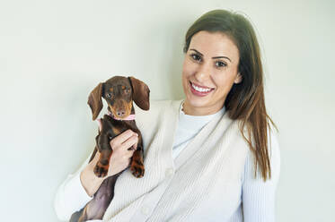 Smiling woman holding dog while leaning on wall at home - KIJF03610