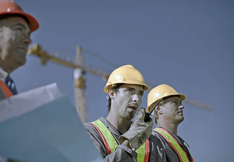 Male construction worker talking on walkie-talkie by coworkers at construction site against clear sky - AJOF01072