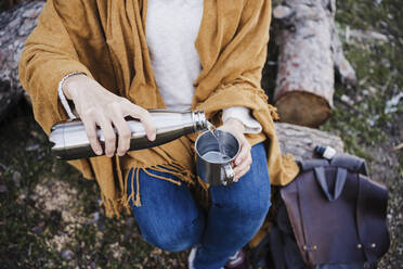 Woman pouring water in cup from thermos while sitting on tree trunk at forest - EBBF02479