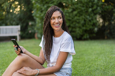 Smiling braided woman with mobile phone looking away while sitting in garden during summer - AKLF00073