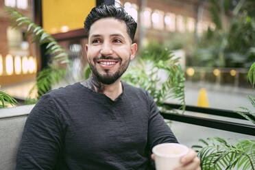 Lächelnder Hipster-Mann mit Kaffeetasse, der wegschaut, während er in einem Straßencafé sitzt - JCCMF01243