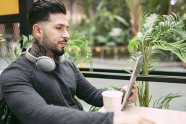 Gutaussehender Hipster-Mann mit Kopfhörern, der ein digitales Tablet am Tisch in einem Café benutzt - JCCMF01244