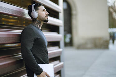 Hipster man with headphones holding digital tablet while standing against metal wall - JCCMF01250