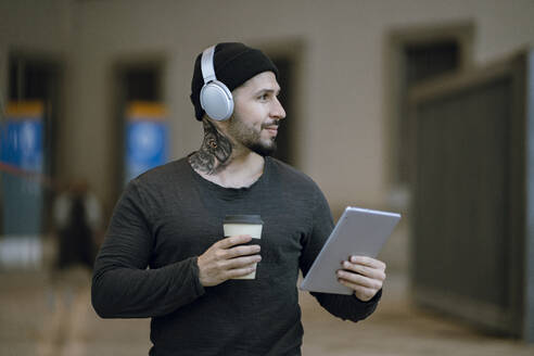 Lächelnder Hipster-Mann mit Kaffeetasse, der ein digitales Tablet hält und wegschaut - JCCMF01252