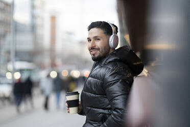 Smiling hipster man with wireless headphones holding coffee cup while looking away - JCCMF01255