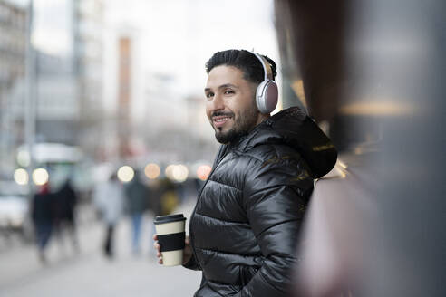 Lächelnder Hipster-Mann mit drahtlosen Kopfhörern, der eine Kaffeetasse hält und wegschaut - JCCMF01255