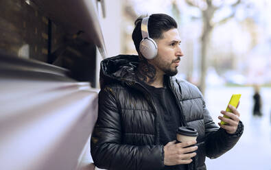 Hipster man with headphones holding coffee cup and smart phone while looking away - JCCMF01257