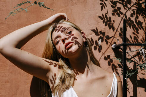 Junge Frau gegen die Wand mit Blättern Schatten auf dem Gesicht während des sonnigen Tages - TCEF01526