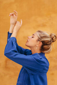 Beautiful blond woman dancing with hands raised in front of yellow wall - TCEF01535