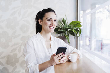 Lächelnde Geschäftsfrau mit Mobiltelefon, die durch ein Fenster im Büro schaut - EBBF02486