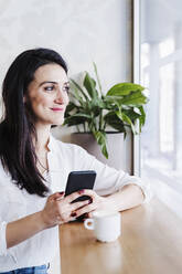 Beautiful woman looking through window while using mobile phone in office - EBBF02489