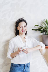 Happy woman holding coffee cup while standing against wall - EBBF02490