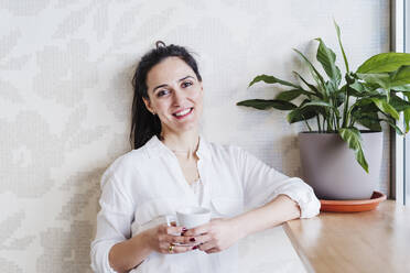 Smiling beautiful woman holding coffee cup while standing at desk against in office - EBBF02494