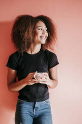 Glückliche Afro-Frau, die eine Kaffeetasse hält und vor einem pfirsichfarbenen Hintergrund wegschaut - EBBF02497