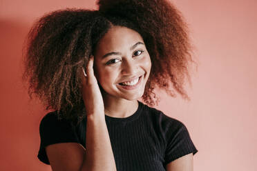 Glückliche Afro-Frau mit Hand im Haar vor pfirsichfarbenem Hintergrund - EBBF02499