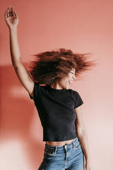 Sorglose Afro-Frau mit erhobenen Armen tanzt vor pfirsichfarbenem Hintergrund - EBBF02501