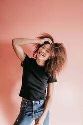 Fröhliche Afro-Frau mit Hand im Haar vor pfirsichfarbenem Hintergrund - EBBF02502