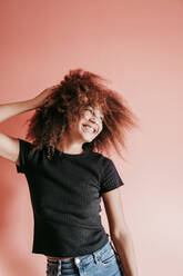 Fröhliche Afro-Frau mit Hand im Haar, die vor einem pfirsichfarbenen Hintergrund wegschaut - EBBF02503